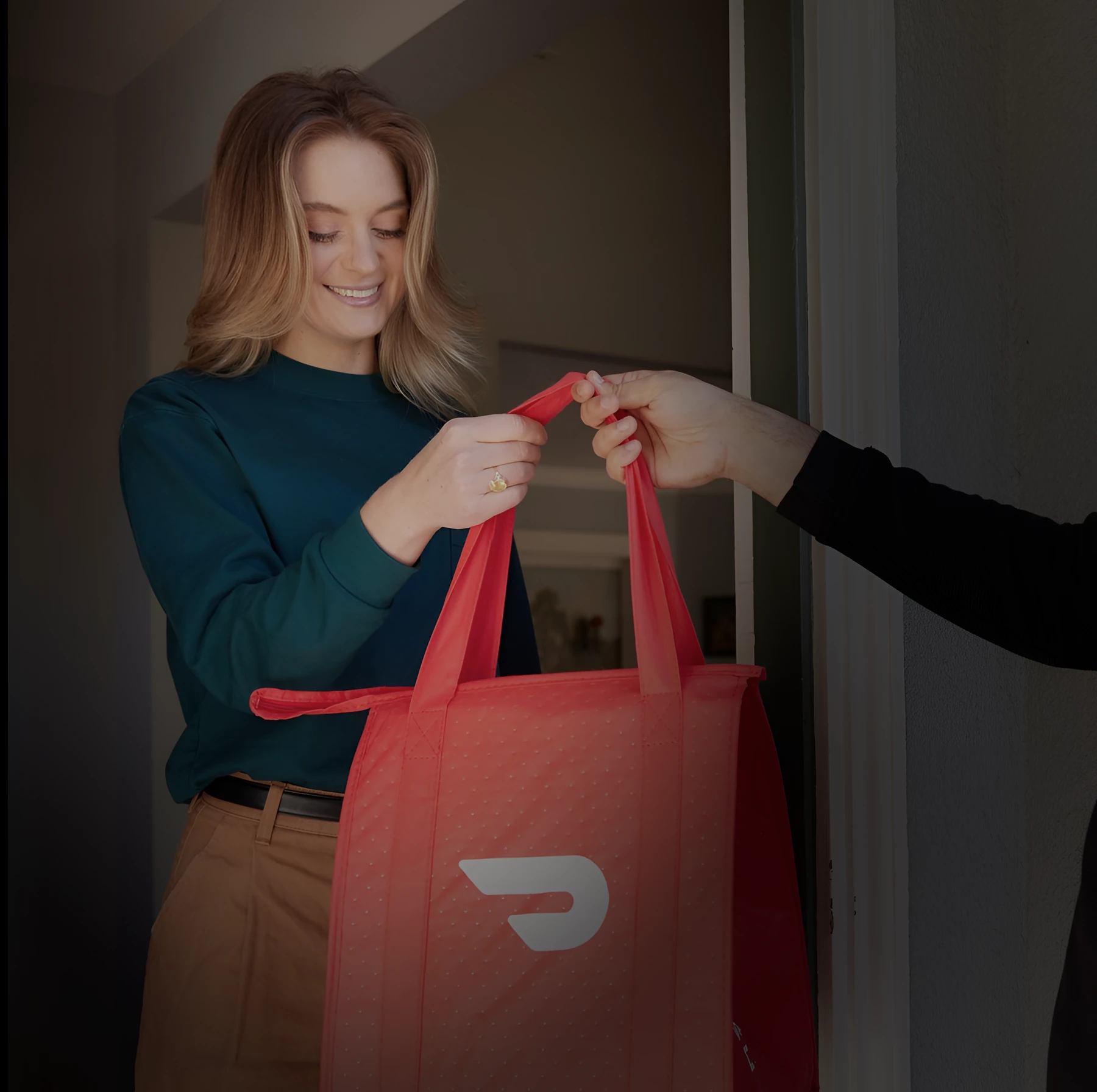 A courier hands a red DoorDash bag to a customer at her front door.